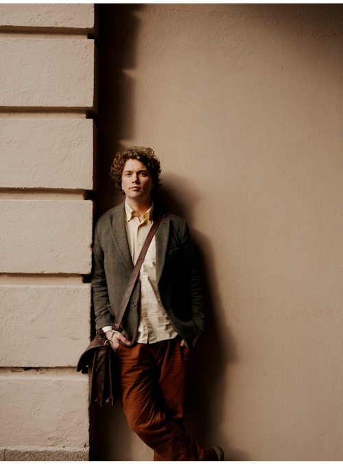 Foto av Kristian Hoff-Andersen ved Ulvetrappa. Han har på seg en brun dressjakke og burgunderfargede bukser. Han lener seg mot veggen.