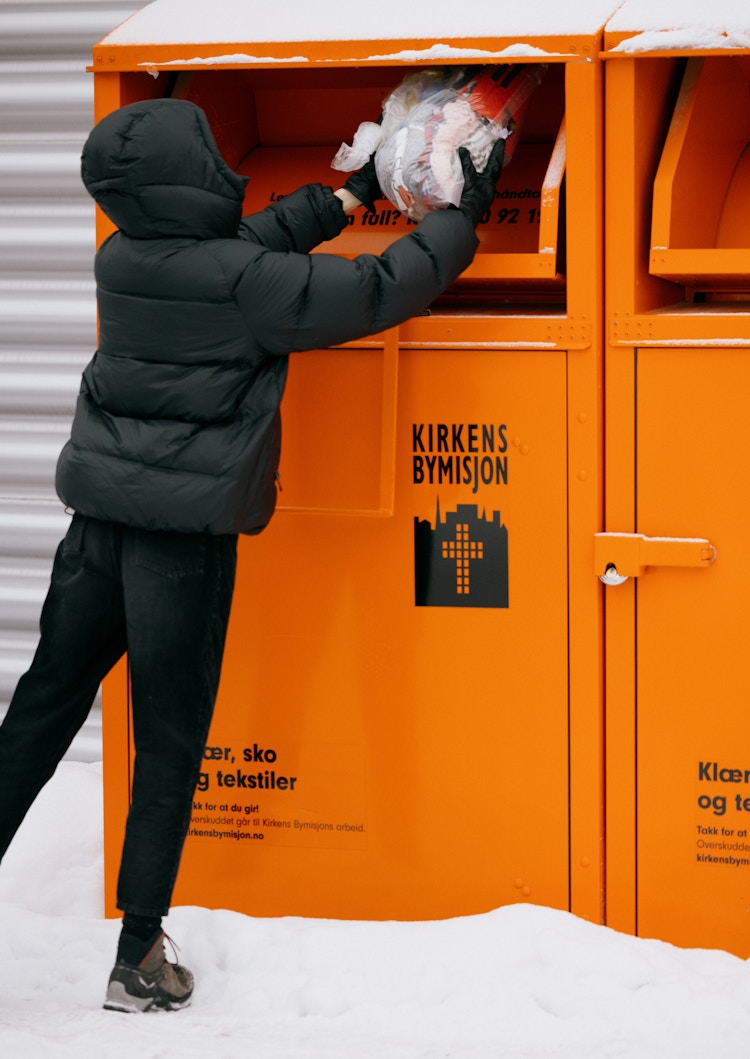 Foto av en person i sort dunjakke som kaster en poste med tøy i Kirkens Bymisjon sine oransje innsamlingsbokser.