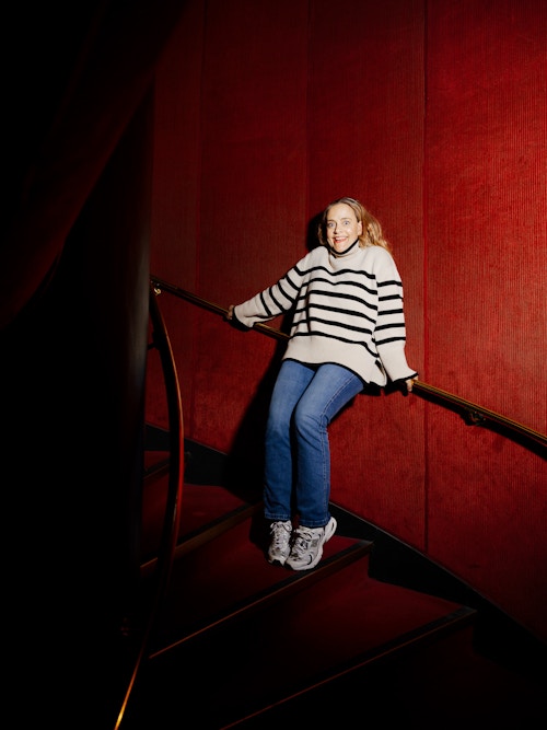 Foto av Henriette Steenstrup i en rødtrapp, hun har hoppet opp på rekkverket og lager en grimase.