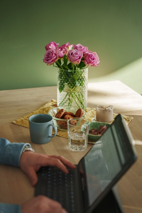 En mann sitter med ipaden på kjøkkenbordet. Veggen er grønnmalt. Det er en vase med rosa blomster i, en kaffekopp, kjeks og vann.
