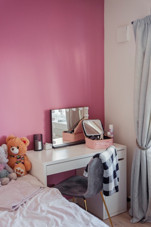 Et barnerom med rosa vegg. Det står en skrivepult der med et speil med lys og en sminkepung. Grå gardiner. Det er et par bamser på senga.