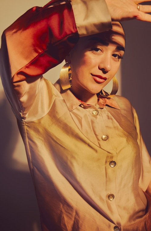 Et portrettbilde av Amanda Delara. Hun har på seg en silkeskjorte i gull. Hun holder armen opp over hodet og titter inn i kamera.