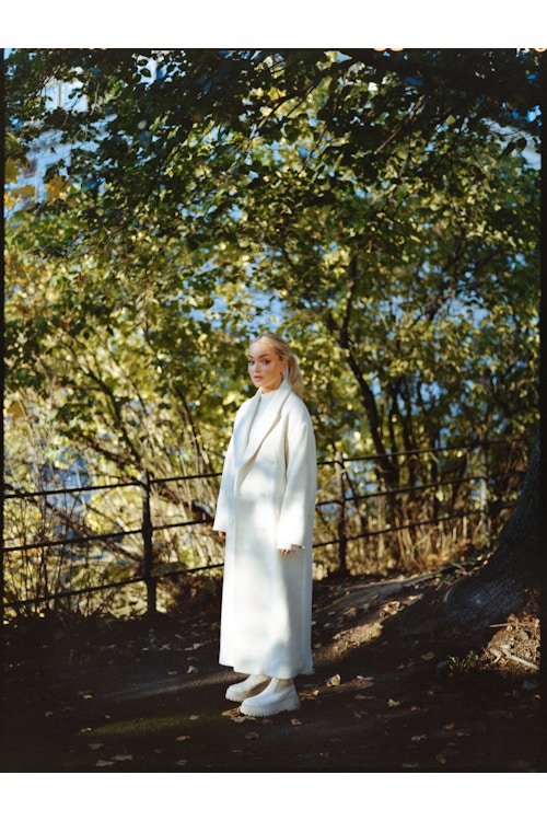 Sophie Elise under noen trær, hvor hun er ikledd hvit kjole, hvit kåpe og hvite sko.