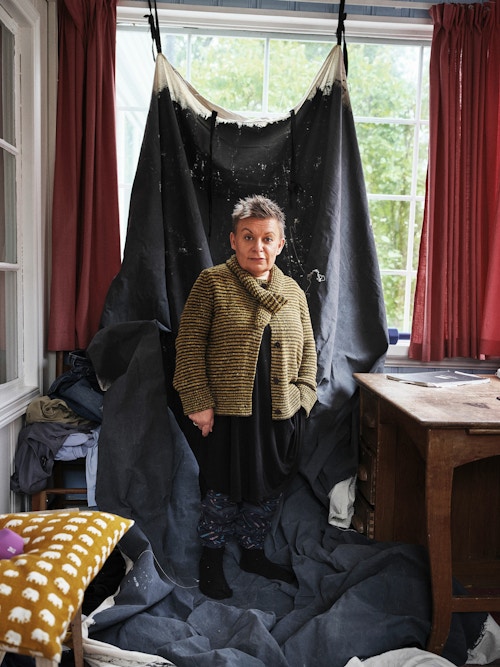 Foto av Kjersti Horn som står hjemme i stua på et teppe som henger oppetter vinduet.