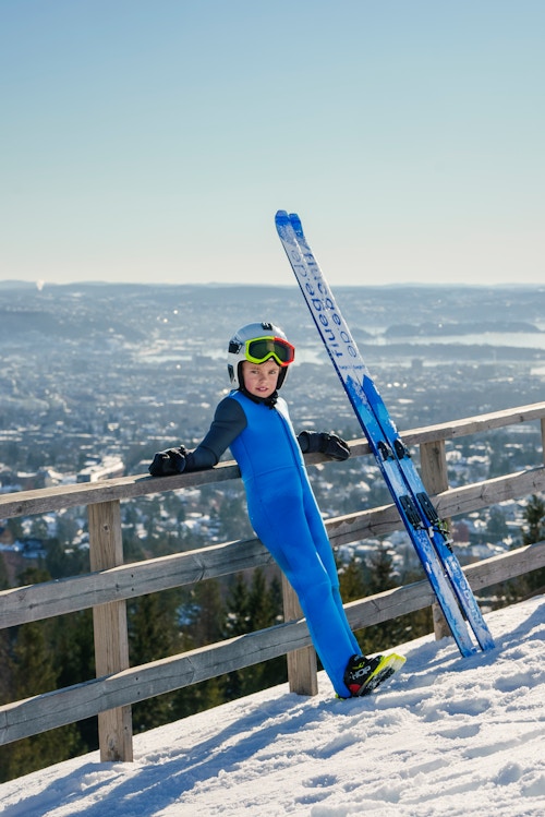 En unge i blå skidress lener seg mot et gjerde med utsikt utover Oslo-fjorden i bakgrunnen.