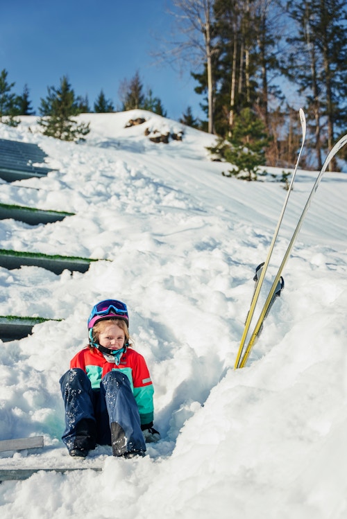 Jente sitter i snøen og myser ved ett av trinnene opp mot toppen av hoppbakken.
