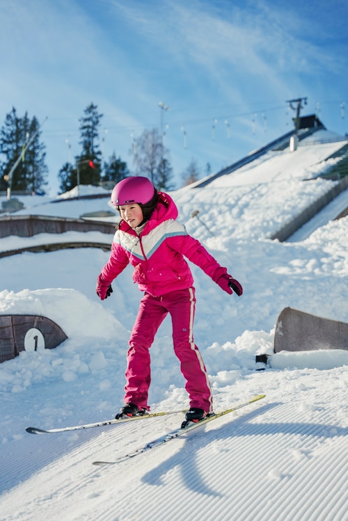 Jente i rosa dress lander etter et lite skihopp. Det er sol og snø