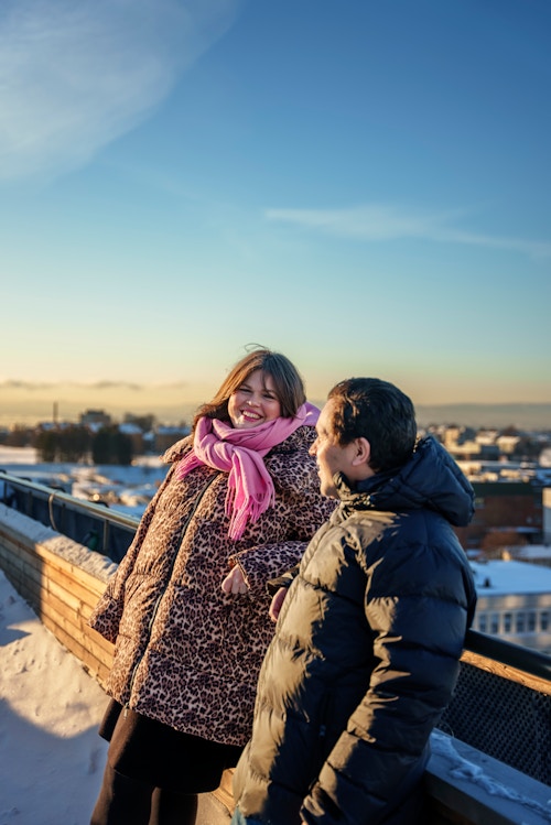 En kvinne og en mann som ser mot hverandre og ler ute på en takterrasse på vinteren