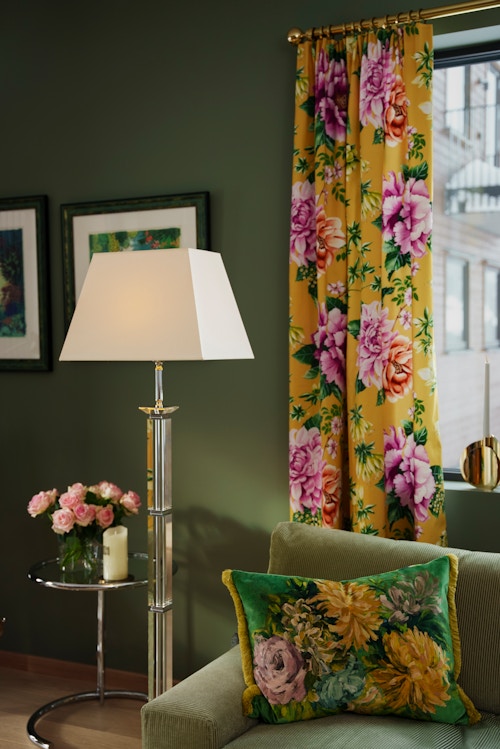 Detaljbilde som viser deler av sofaen, gardinene og lampen i stuen. Veggen og sofaen er grønn, lampen er hvit og gardinene er gule med rosa blomster.