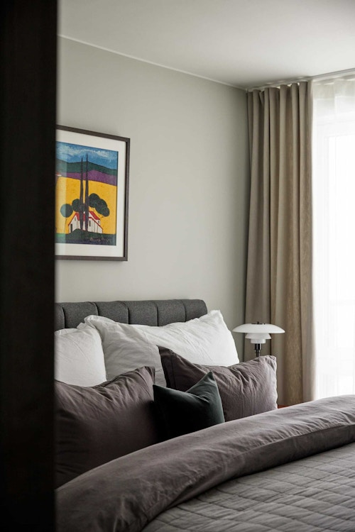 Bilde av et gråmalt soverom ved beige gardiner, grå dobbeltseng og et fargerikt bilde på veggen.