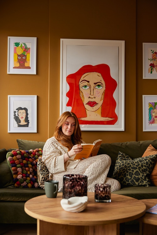 Martine sitter i sofaen sin og leser en bok. Veggene som er malt i oransje er dekorert med flere illustrasjoner hun har tegnet selv.