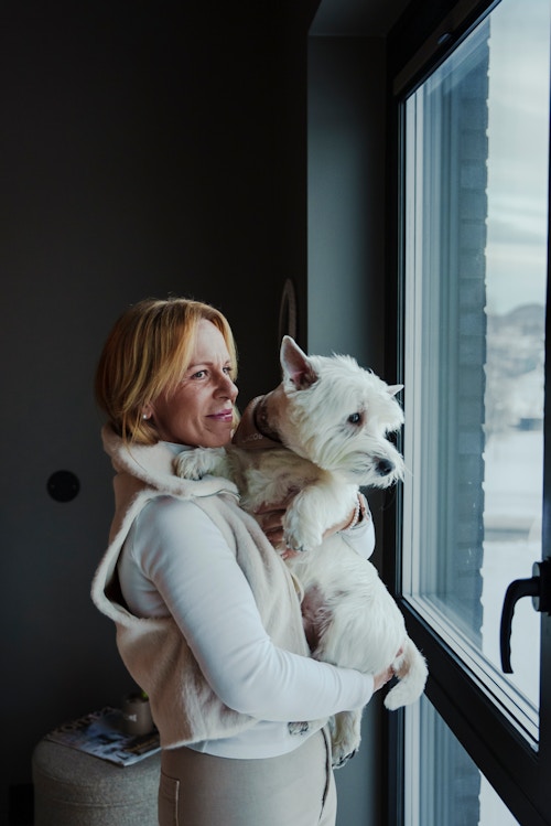 Nærbilde av Ann-Christin som holder en hund og ser ut mot utsikten fra et soverom
