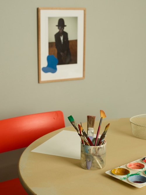 Et rundt bord med malesaker. En rød stol ved siden av og et bilde på veggen.