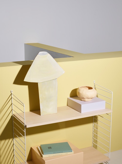 En hylle fra String hengt opp på en gul vegg, med bokser, bøker og en skål plassert i hyllene. Øverst står en figur i lampeform, skjært ut i papp.