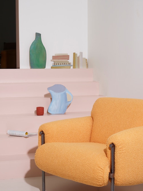 En gul lenestol foran en rosa trapp. I trappen står det ulike objekter laget i papp, som en kanne, en flaske og bøker.