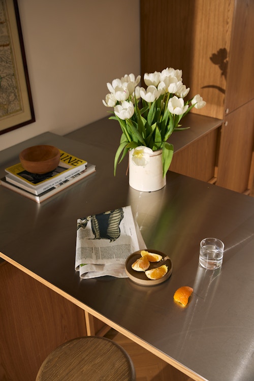 Bilde av bordplaten på barløsningen. Her ligger det også en avis og et fat med oppskåret appeilsin. Det er også en vase med blomster på bordet.