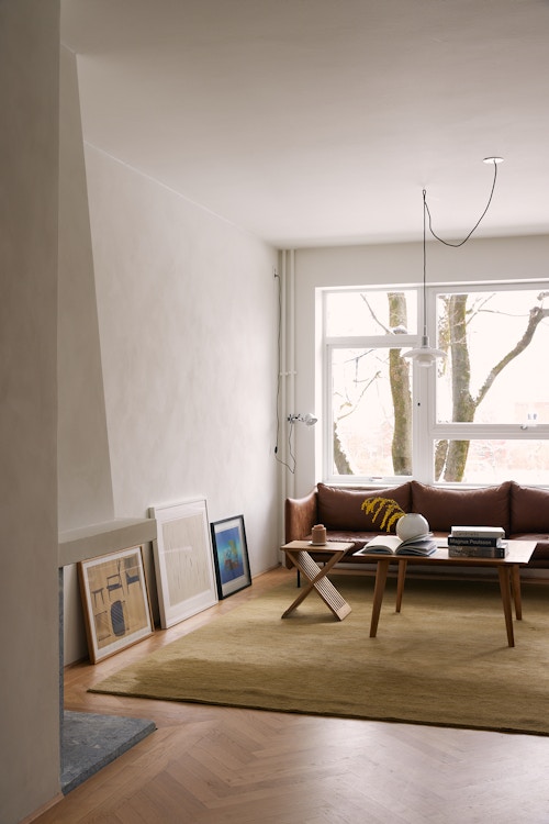 Bilde av stuen med sofa, teppe, bilder som står langs gulvkanten og et stort vindu på den ene veggen.