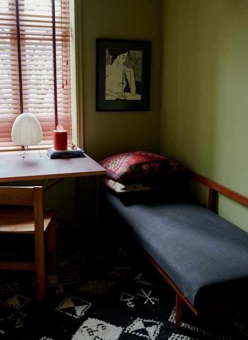 Et skrivebord og en liten seng foran et vindu i et rom med grønne vegger.