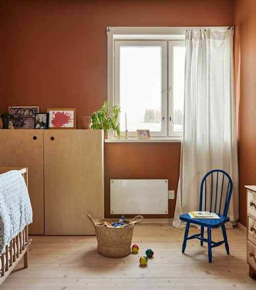 Oversiktsbilde av barnerom malt i en oransje tone, med barneseng, skap, kommode, blå stol, vindu og gardiner.