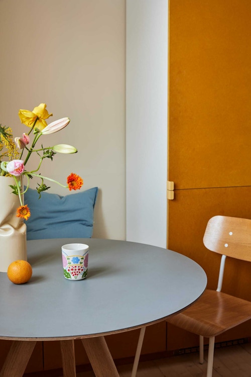 Kjøkkenbord med blomster, kaffekopp og en appelsin. En spisestuestol står foran et gult kjøkkenskap.