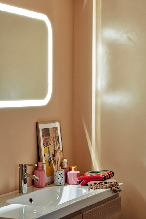 Bildet viser et hjørne av baderommet, med mikrosement i en varm farge på veggene, speil med lys og servantskap.