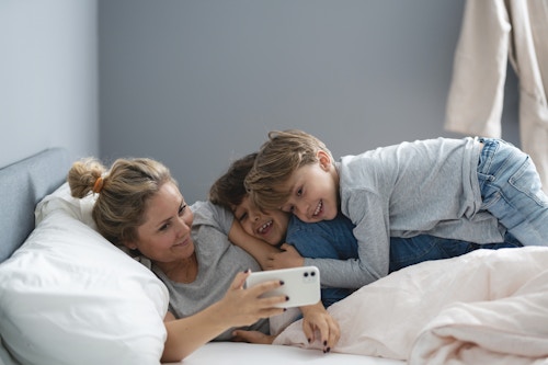 Mor og to barn ligger i senga og ser på noe på mobilen til mor.