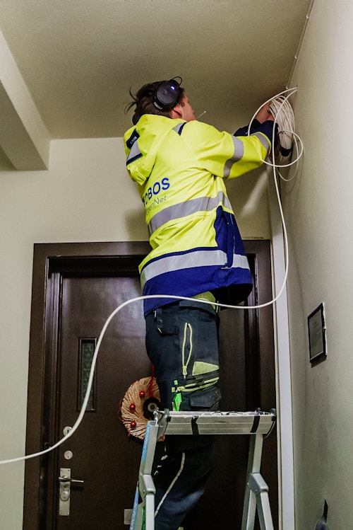 En tekniker står på en stige og forer fiberkabel gjennom veggen til en leilighet.