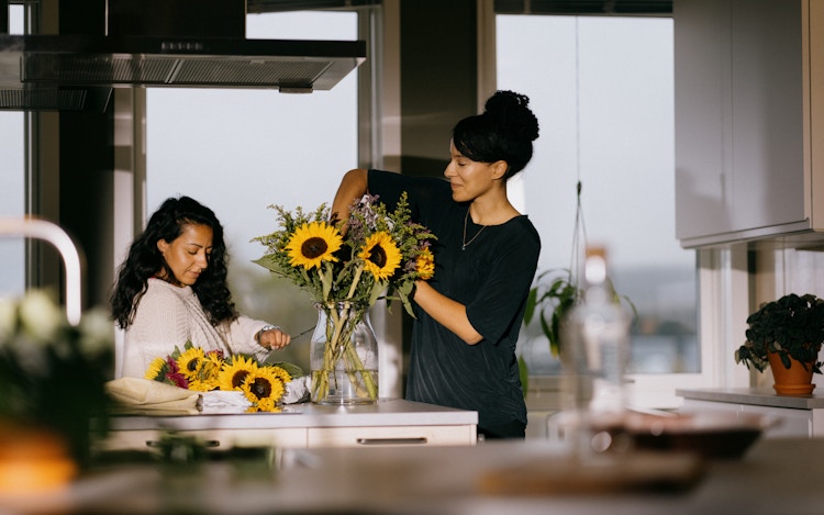 Foto av to kvinner på et kjøkken som arrangerer blomster i en vase.