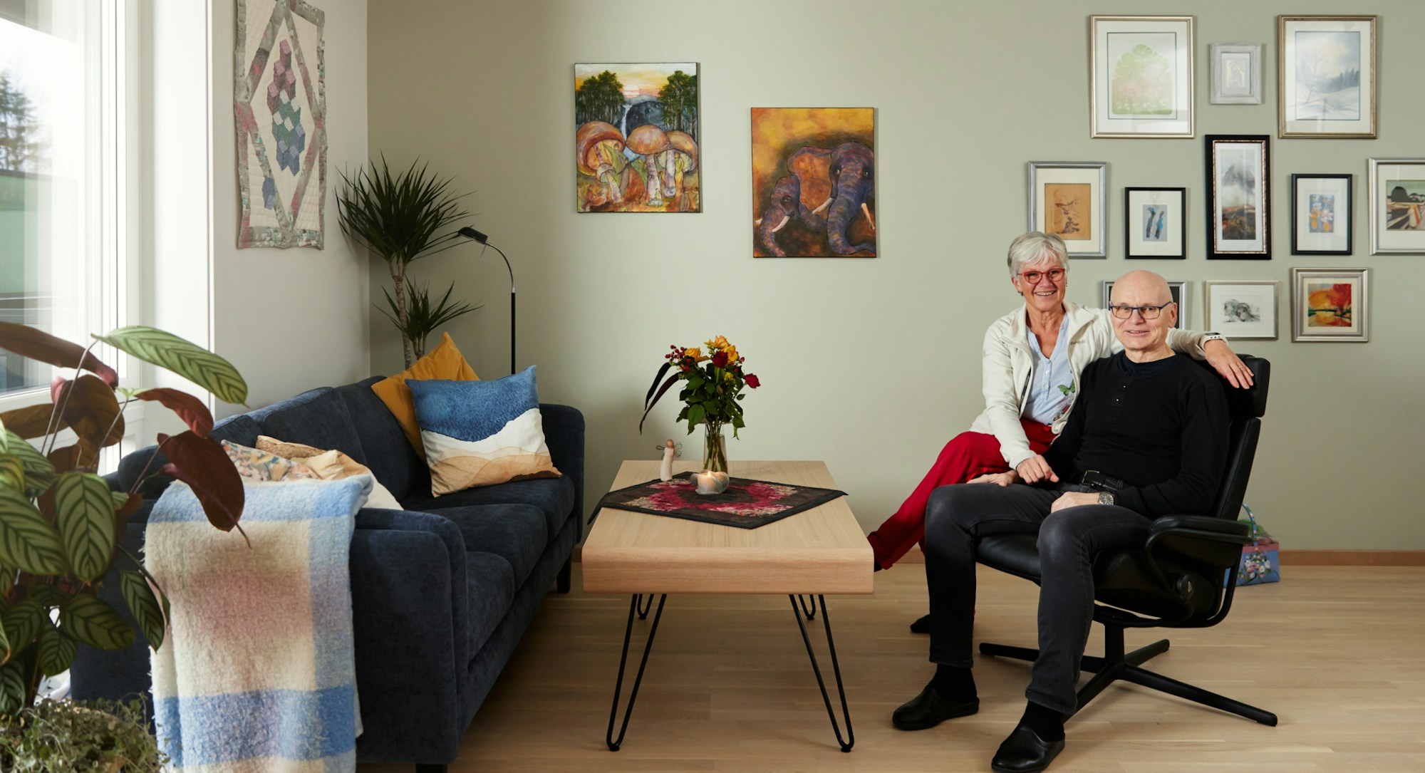Foto av Solveig og Per som sitter i stua i leiligheten sin, med mange bilder på veggene.