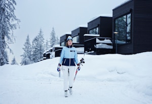 En dame går med skiene sine nedover bakken foran huset sitt i Vitigrend, hun har på seg skiutstyr med skiforeningens logo.
