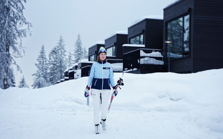En dame går med skiene sine nedover bakken foran huset sitt i Vitigrend, hun har på seg skiutstyr med skiforeningens logo.