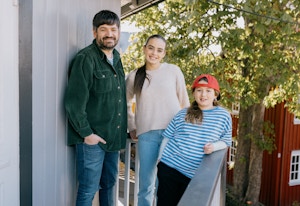 Tre personer lener seg mot et rekkverk ved et hus. En mann står i grønt og smiler. To jenter står ved siden av, en med caps.