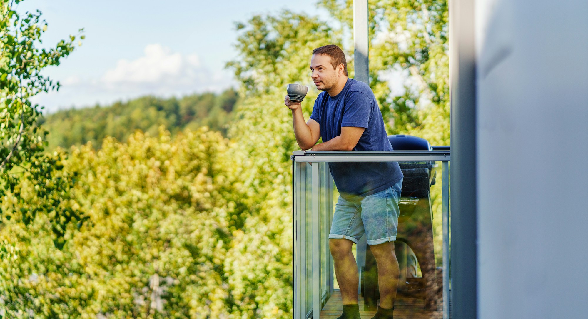 Aleksei står på balkongen sin med en kaffekopp i hånda og ser på utsikten.